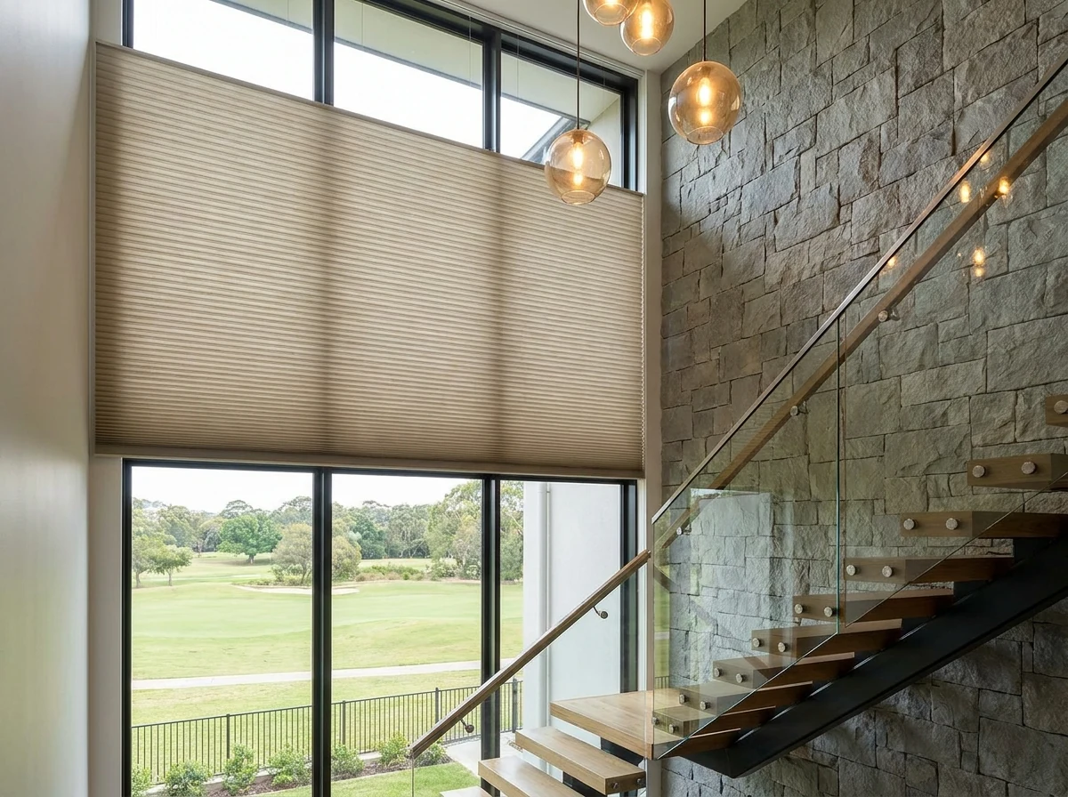 Double-volume glazed stairwell in a Modern Highveld estate home, a cellular honeycomb blind with a visible pleated cell structure lowered across the upper half of tall glazing, open below to a golf fairway view, warm pendant lighting against a stone feature wall